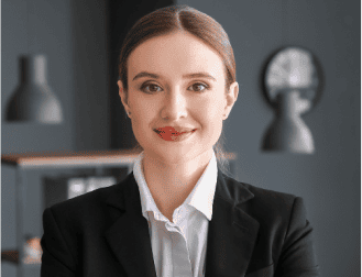 A trusted Chicago personal injury lawyer with brown hair tied back, wearing a black blazer and white shirt, stands indoors in an office setting, smiling at the camera.