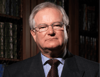 An older man with gray hair and glasses, wearing a suit and tie, stands in front of bookshelves—embodying the experience and professionalism of a Trusted Chicago Personal Injury Lawyer.