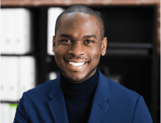 A trusted Chicago personal injury lawyer in a dark blue blazer and black shirt smiles at the camera, with shelves and binders blurred in the background.