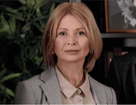 A woman with blonde hair in a bob hairstyle, wearing a light gray blazer and beige shirt, stands indoors with green plants and framed photos in the background—an atmosphere reminiscent of About Lawyer Furqan’s professional setting.