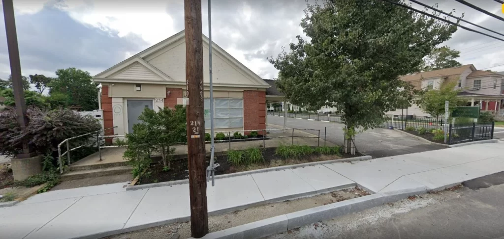 A small brick building with a ramp and stairs at the entrance, surrounded by greenery, next to a paved sidewalk and street, where Founder Furqan Mohammed speaks at a webinar.