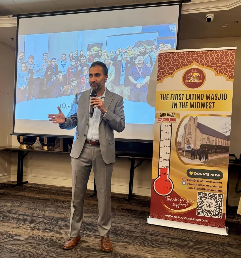 Founder Furqan Mohammed speaks into a microphone next to a fundraising banner and a projected group photo at an indoor event.