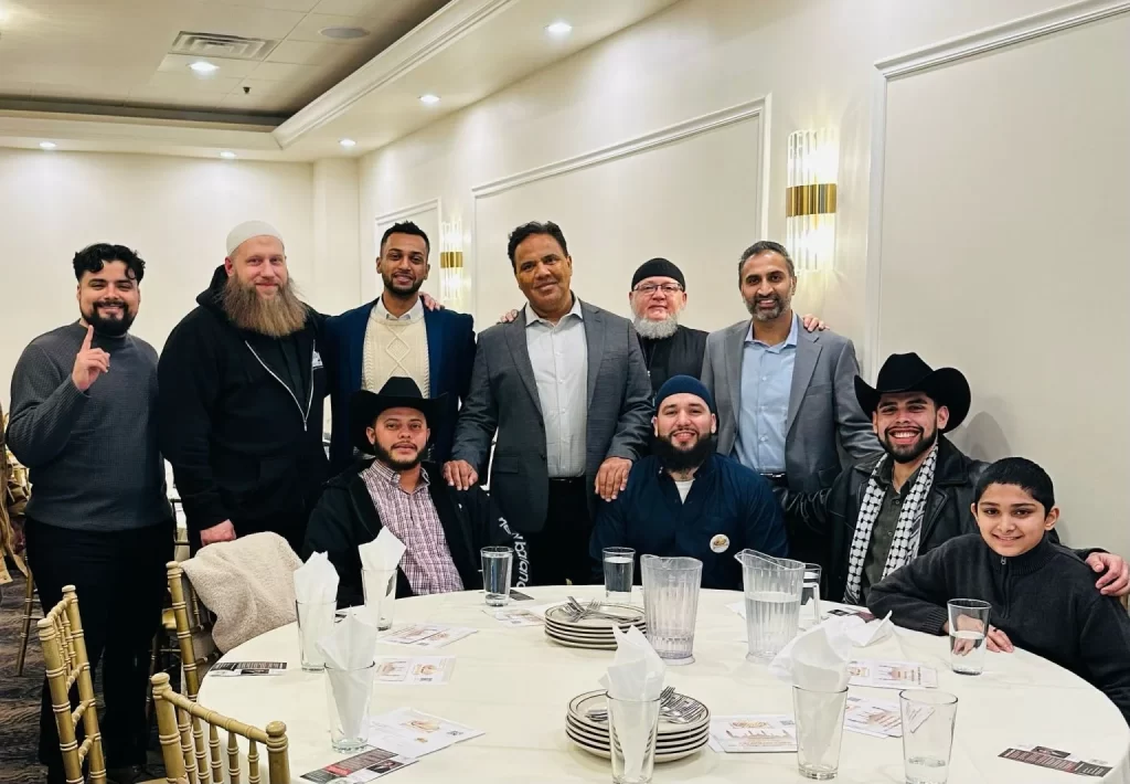 A group of eleven people, some seated and some standing, pose for a photo around a round table set for a meal in a banquet hall, shortly after Founder Furqan Mohammed speaks at the webinar.
