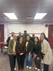 A group of ten people stand and smile in a classroom in front of a whiteboard filled with handwritten notes after Founder Furqan Mohammed speaks at webinar.