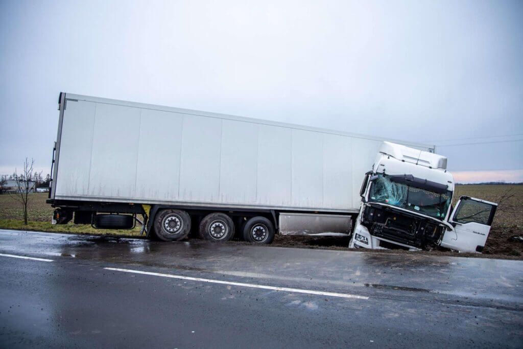 A large white semi-truck is off the road, with its front cab damaged and tilted into a muddy ditch beside a wet highway.