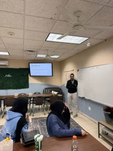 A classroom with three students seated at a table and a teacher standing near a whiteboard. A presentation titled "Negligence Law" is displayed on a screen for the Injury & Car Accident Seminar.