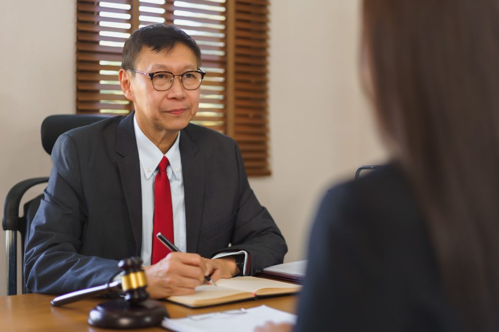 A man in a suit sits at a desk with a notebook and gavel, listening to a woman sitting across from him in an office setting.
