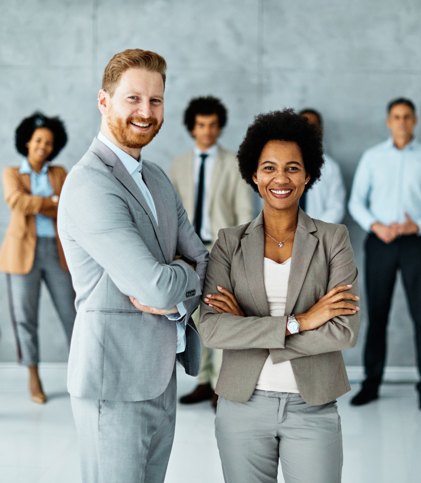 Two professionals in business attire stand confidently with arms crossed in front of a group of colleagues against a modern, gray office background.