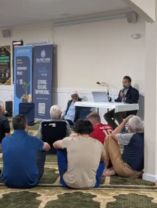 Furqan and Amro speak at a Boston Islamic Estate Planning Seminar, addressing an audience seated on the floor in a community hall, with informational banners and a laptop on the table beside them.