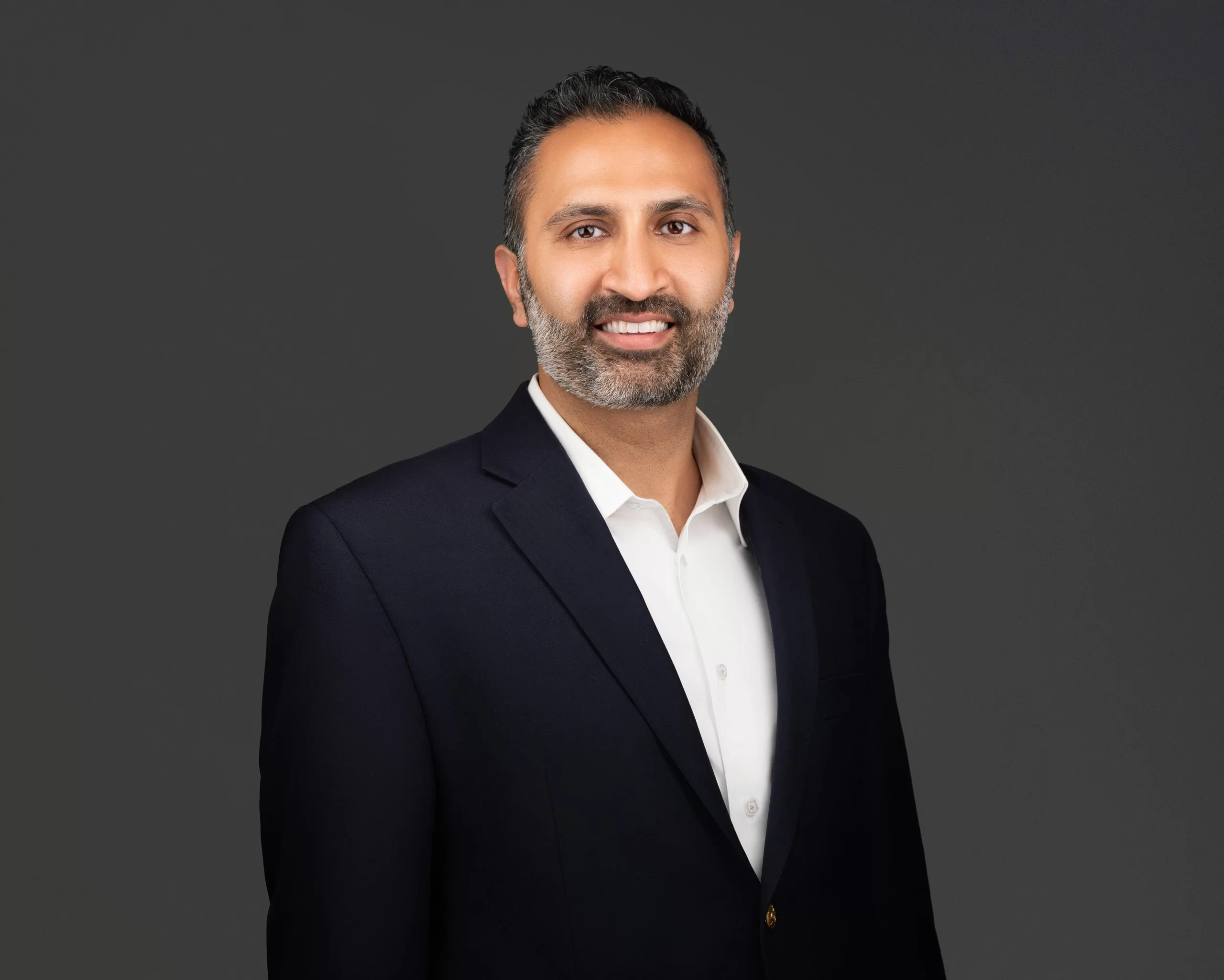 Man with short dark hair and beard, trusted Chicago personal injury lawyer, wearing a navy suit jacket and white shirt, smiling at the camera against a plain dark background.
