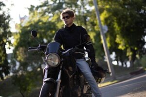 motorcyclist wearing jacket and sunglasses seated on motorcycle near tree-lined road