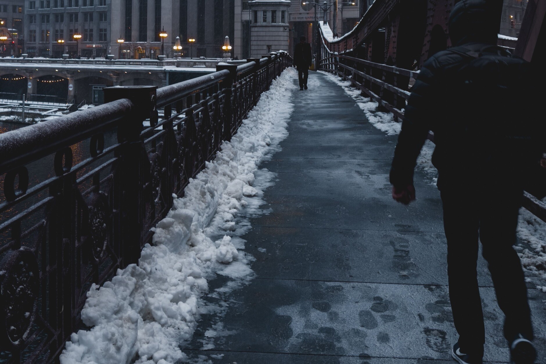 Cleared walkway on a Chicago bridge with snow piled along both edges and a wet, slick surface underfoot