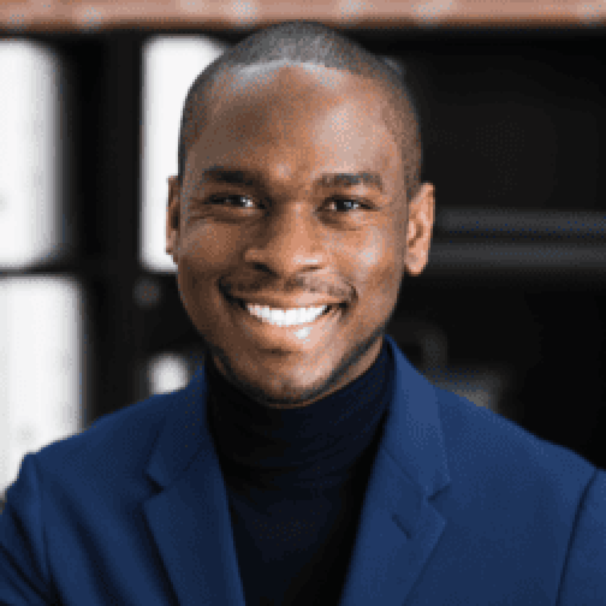 A trusted Chicago personal injury lawyer in a dark blue blazer and black shirt smiles at the camera, with shelves and binders blurred in the background.